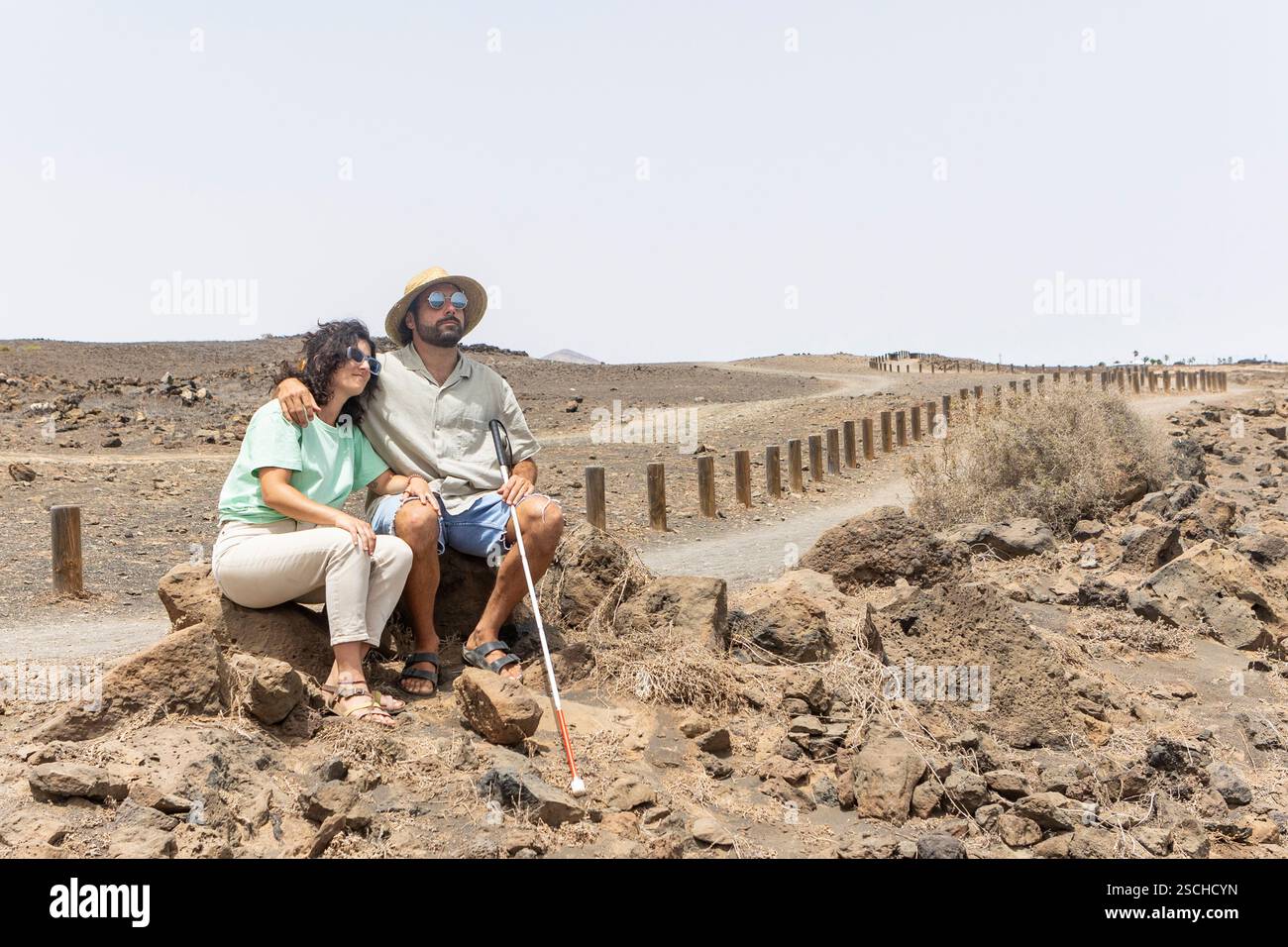 Un aveugle avec une canne et une femme assis ensemble sur des rochers le long d'un chemin désertique tranquille. La scène capture un moment de repos et de compagnie pendant thei Banque D'Images