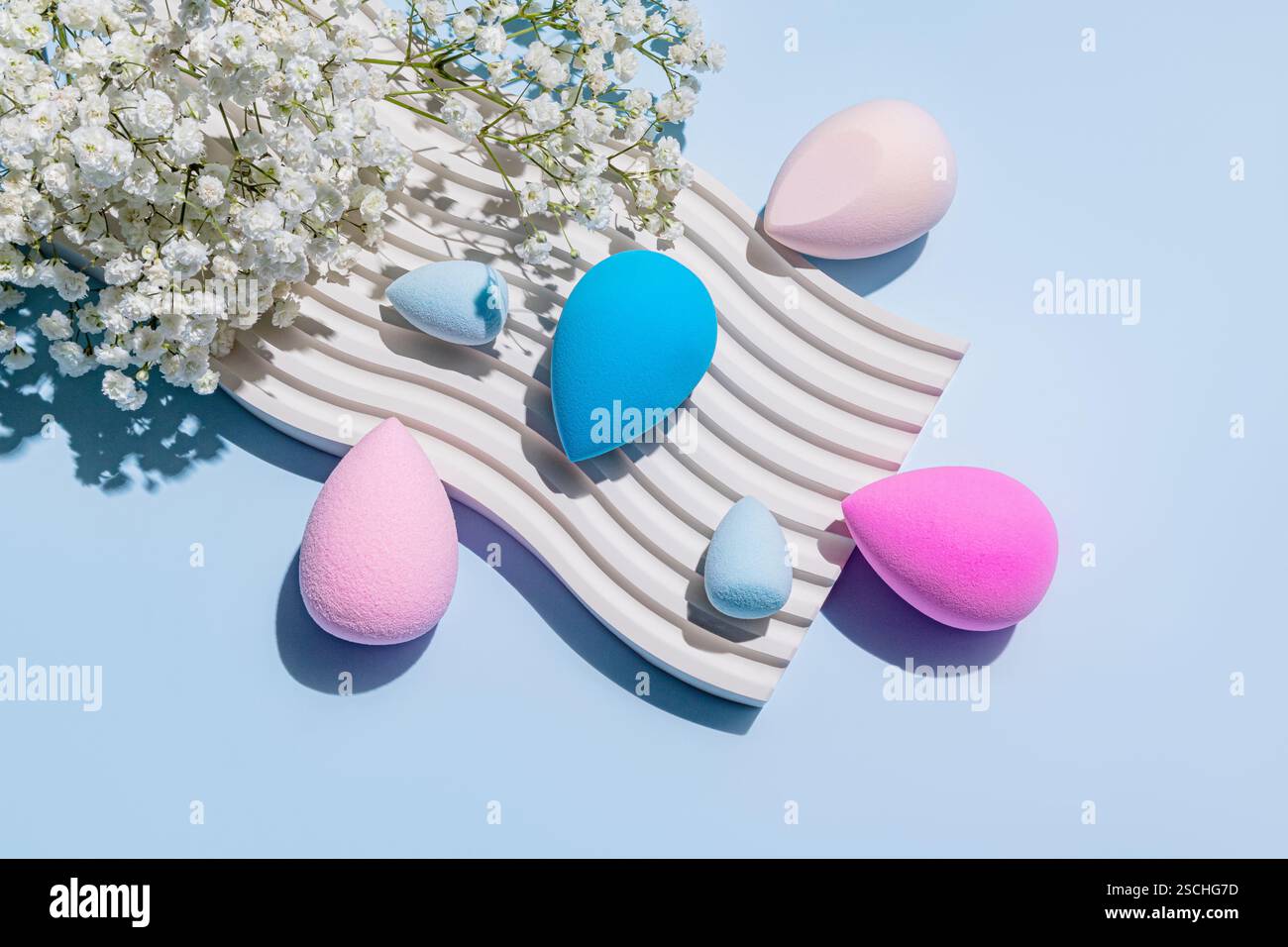 Vue de dessus d'éponges de maquillage vibrantes disposées esthétiquement sur une surface texturée et ondulée à côté de fleurs blanches fraîches, soulignant la beauté et l'hygiène p Banque D'Images