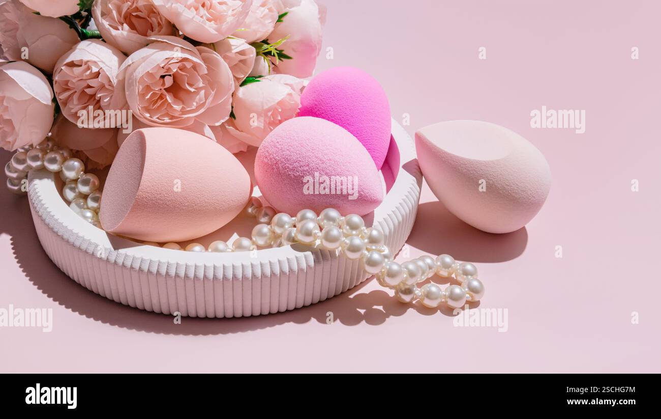 Diverses éponges d'oeufs de maquillage, un mélangeur de beauté, disposés au milieu de fausses perles et de roses roses sur un fond rose clair. Banque D'Images