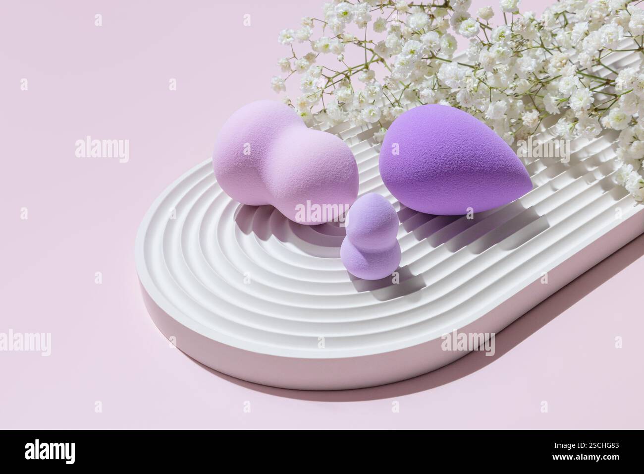 Différentes tailles et formes d'éponges de maquillage aux tons violets habilement mises en scène sur un plateau blanc strié, accompagnées de délicates fleurs blanches sur un dos rose Banque D'Images