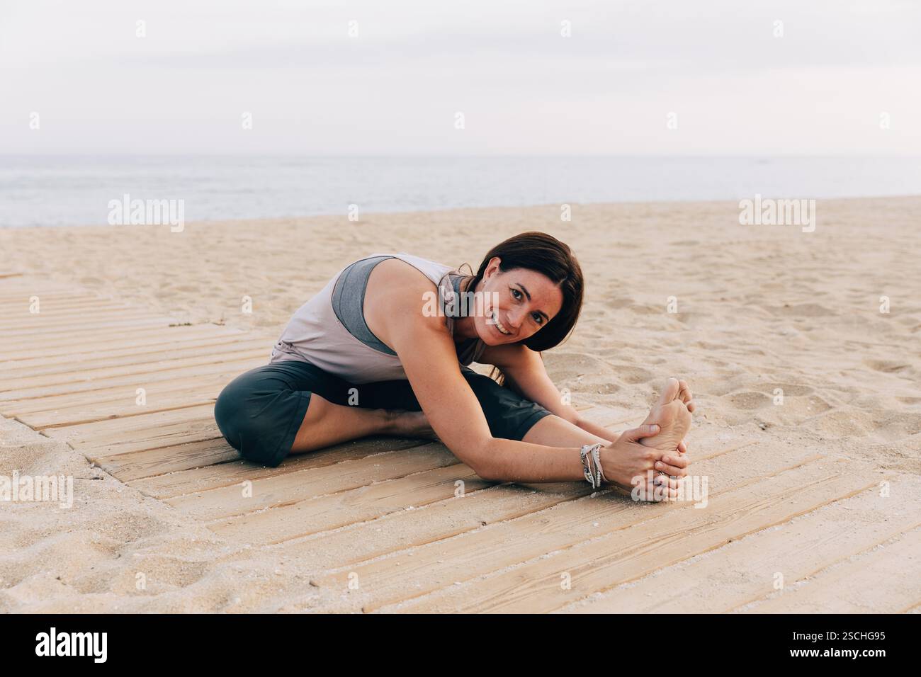 Une femme pratique la pose de yoga Janu Sirsasana sur un tapis de bois à la plage, souriant à la caméra avec l'océan en arrière-plan pendant le coucher du soleil. Banque D'Images