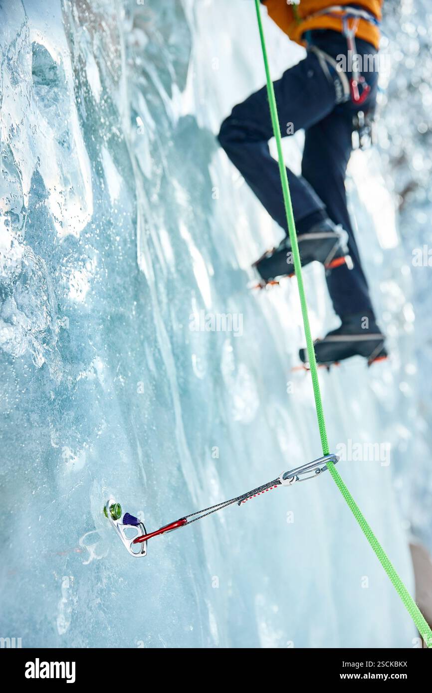 Gros plan mince quickdraw avec mousqueton rouge grimpant accroché les vis à glace annulaires vissées dans la glace d'un mur de glacier gelé contre un athlète grimpeur. Banque D'Images