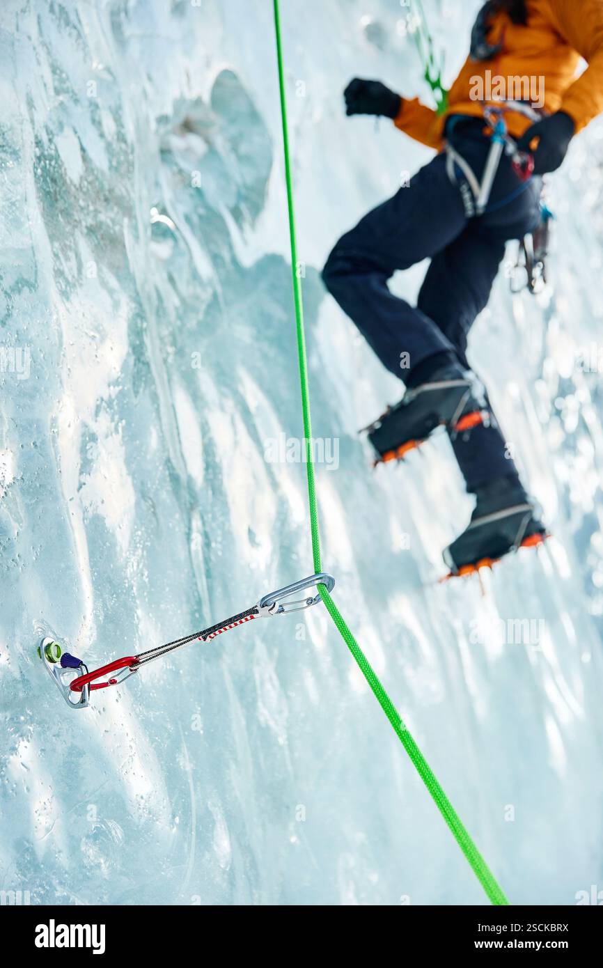 Gros plan mince quickdraw avec mousqueton rouge grimpant accroché les vis à glace annulaires vissées dans la glace d'un mur de glacier gelé contre un athlète grimpeur. Banque D'Images