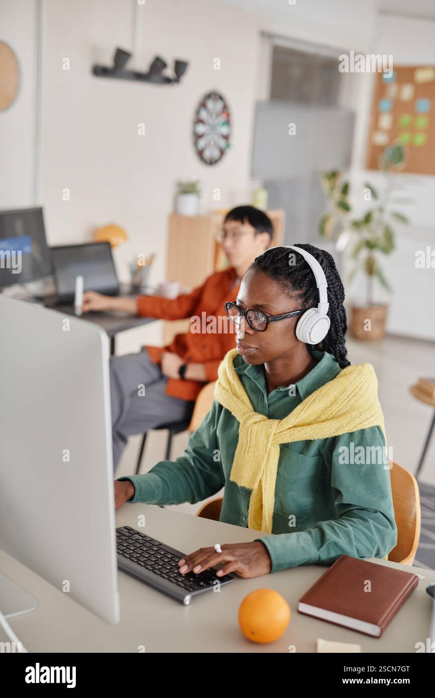 Personne noire travaillant dans un bureau moderne, portant des écouteurs tout en tapant sur le clavier. Collègue en arrière-plan assis au poste de travail, plantes et décoration de bureau présents Banque D'Images