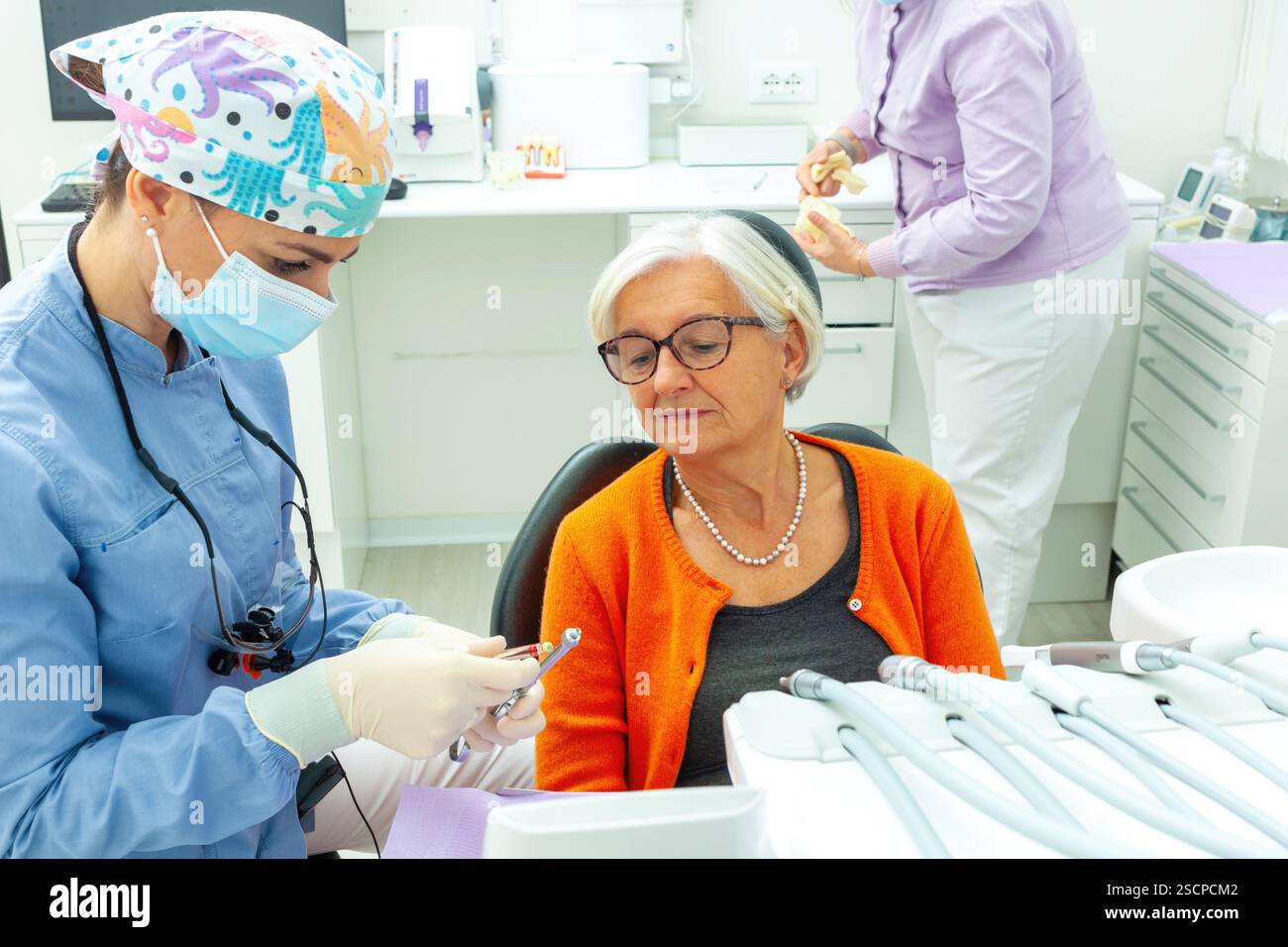Dentiste portant un masque chirurgical et une loupe montrant une perceuse dentaire à un patient senior en clinique dentaire Banque D'Images
