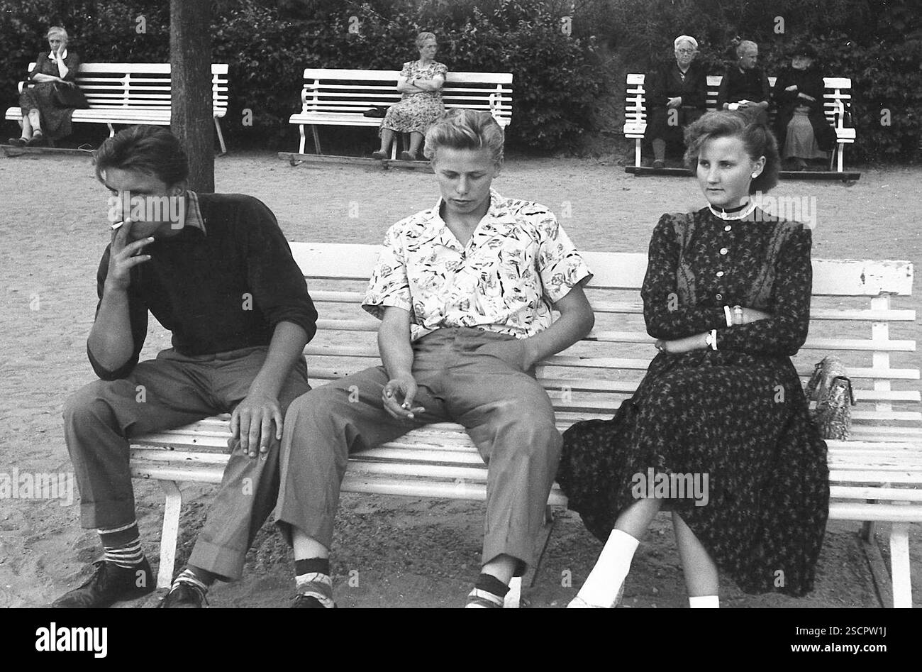 Deux jeunes hommes et une jeune femme en robe sont assis sur un banc du parc sur la promenade de la plage à Ahlbeck. Les deux hommes fument une cigarette. [traduction automatique] Banque D'Images