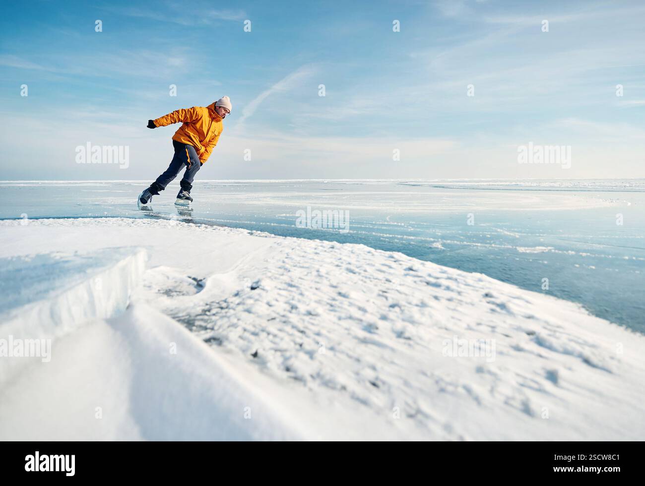 Homme athlète en veste jaune patin sur glace sur le lac gelé avec des fissures et de la neige à l'heure d'hiver Banque D'Images
