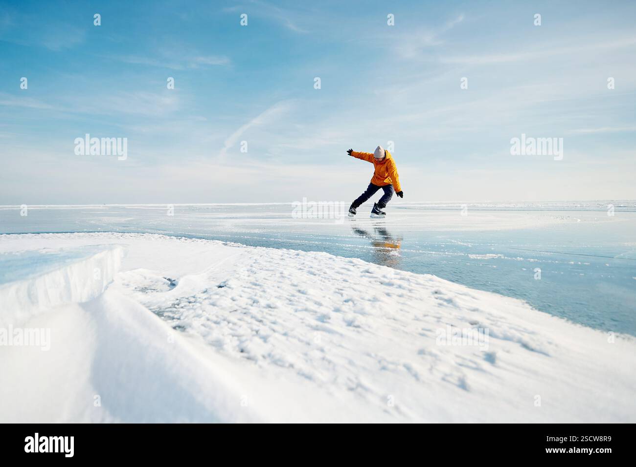 Homme athlète en veste jaune patin sur glace sur le lac gelé avec des fissures et de la neige à l'heure d'hiver Banque D'Images