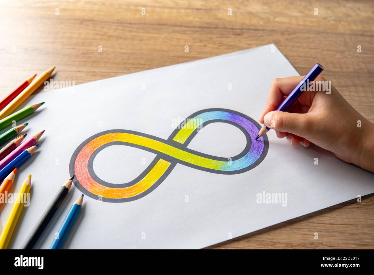 Une main coloriant un symbole d'infini arc-en-ciel sur une feuille de papier blanche avec des crayons de couleur dispersés autour. Concept de sensibilisation à l'autisme. Banque D'Images Une main coloriant un symbole d'infini arc-en-ciel sur une feuille de papier blanche avec des crayons de couleur dispersés autour. Concept de sensibilisation à l'autisme. Banque D'Images