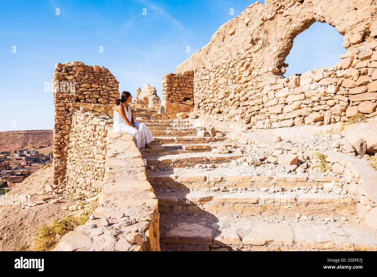 Ait BEN HADDOU, MAROC - 9 nov 2023 : jeune femme en jupe blanche assise sur les marches de pierre du ksar ait Ben Haddou, ancien village berbère en briques adobe ou kas Banque D'Images