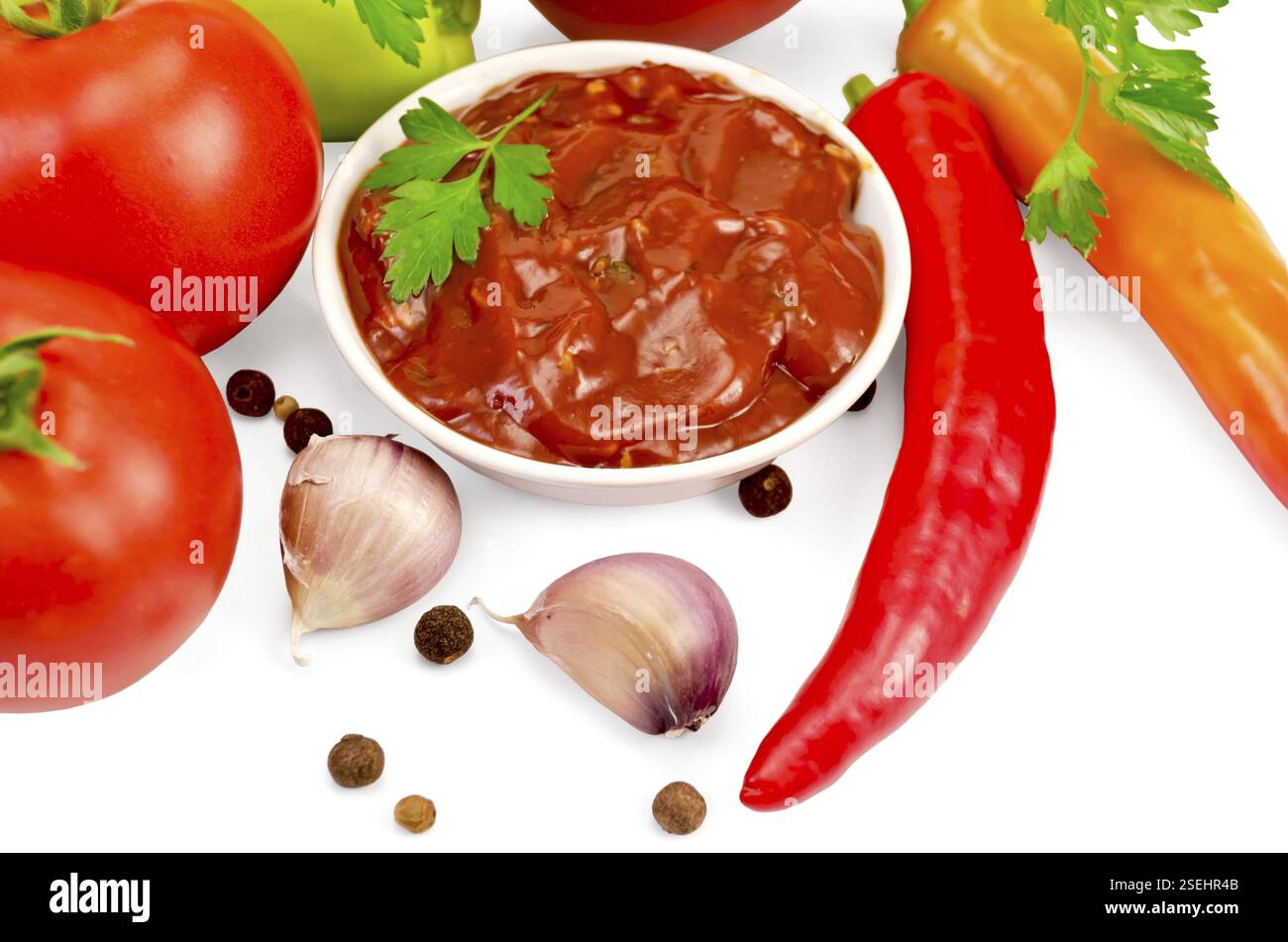 Ketchup dans une tasse blanche, tomates, deux piments rouges frais, ail, grains de poivre, moutarde, persil sur fond blanc Banque D'Images