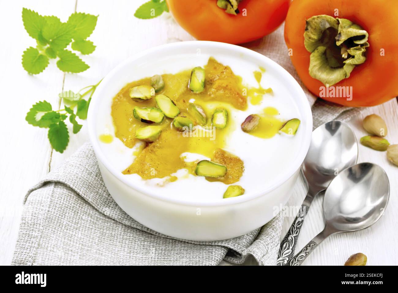 Dessert de yaourt, miel et kaki avec la vanille, la cardamome et pistaches dans un bol sur une serviette, la menthe et la cuillère sur fond de carte en bois clair Banque D'Images