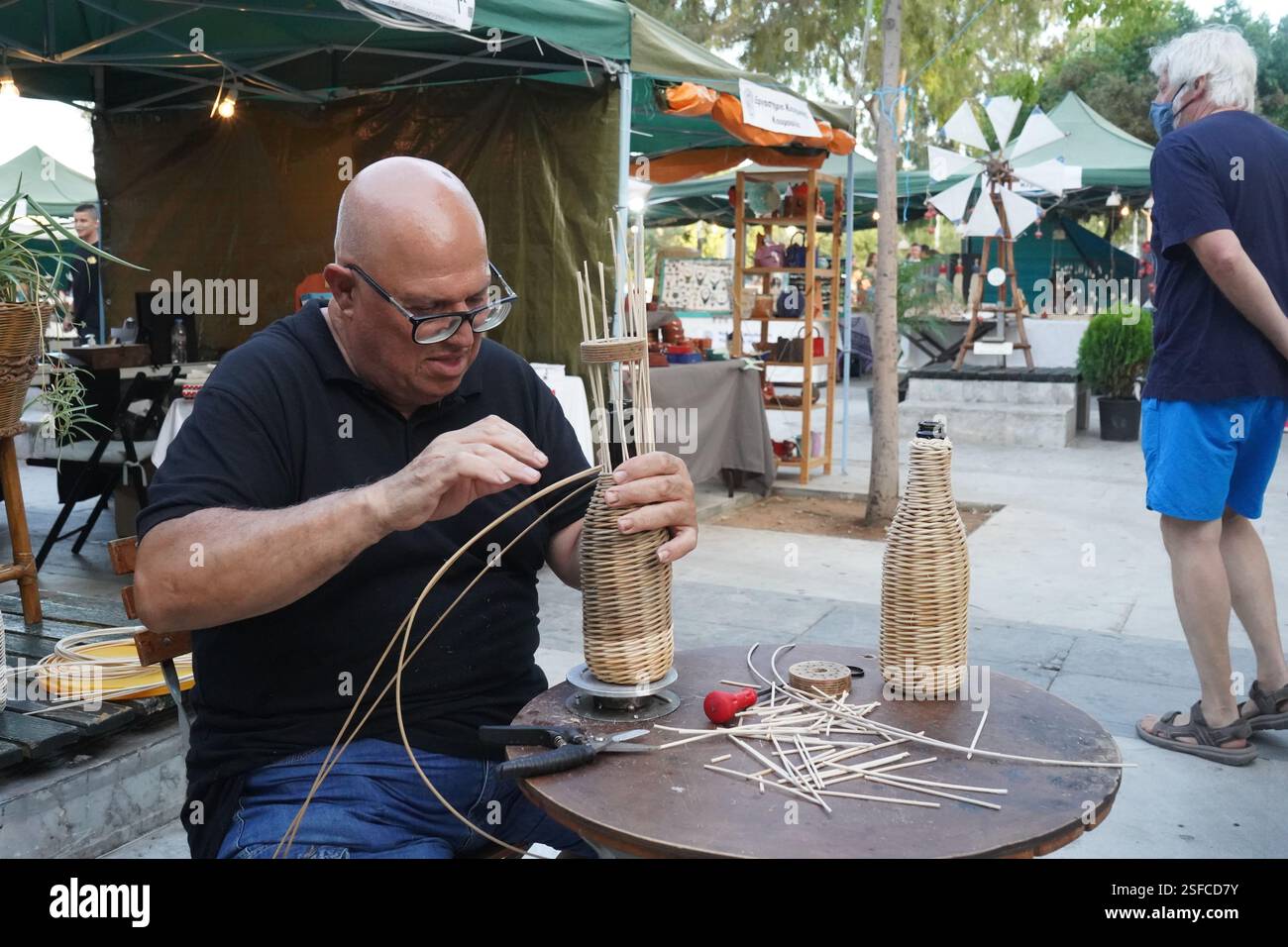 Un homme tisse une bouteille en osier, attachant des tiges de bois autour d'elle. Un espace de travail avec des matériaux prêts pour l'artisanat traditionnel mis sur table Banque D'Images