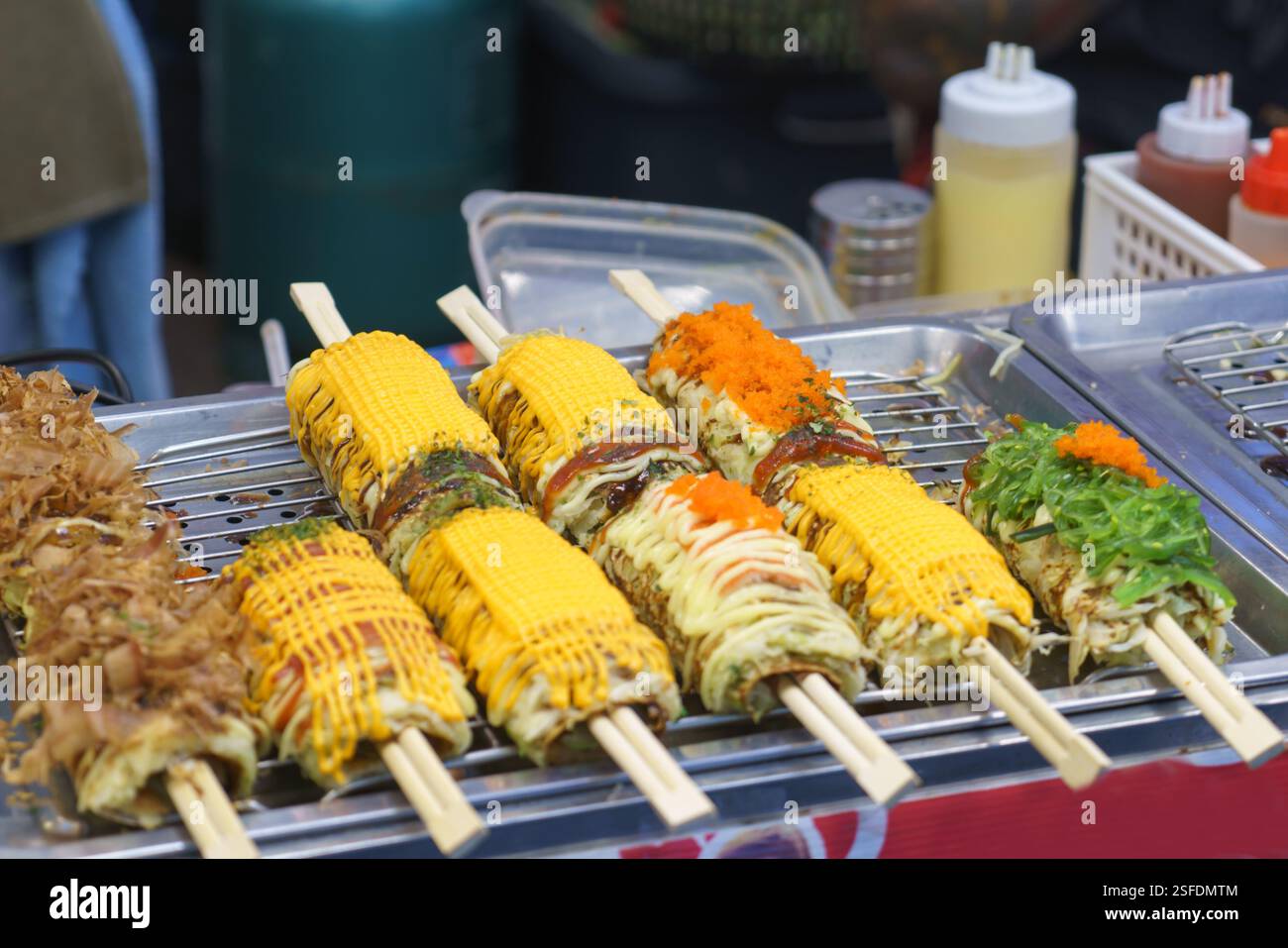 Gros plan de brochettes de maïs grillées de style japonais exposées sur un stand de Street food, ornées de sauces, d'œufs et d'herbes vibrantes. Les pres colorées Banque D'Images