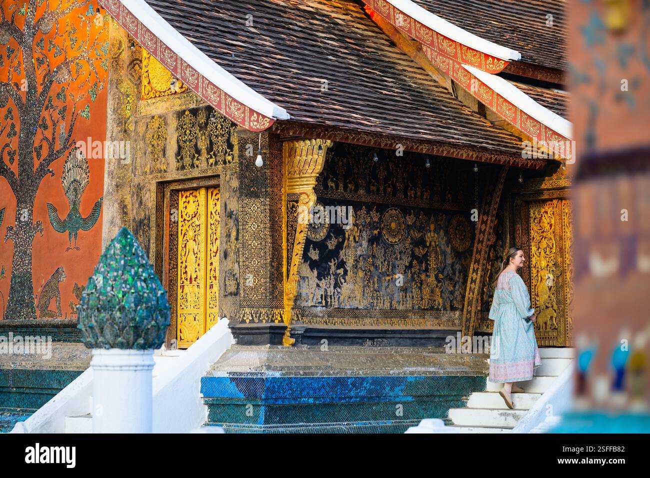 Une femme sur les escaliers du temple Wat Xiengthong à Luang Prabang, au Laos, avec des peintures murales complexes en or et en mosaïque, y compris un arbre décoratif et des animaux Banque D'Images