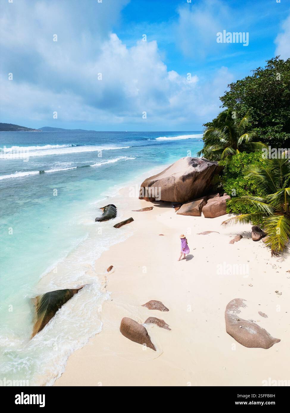 Vue aérienne d'une femme en robe violette se promenant le long d'une plage immaculée sur la Digue, aux Seychelles, avec de grands rochers de granit et des palmiers luxuriants Banque D'Images