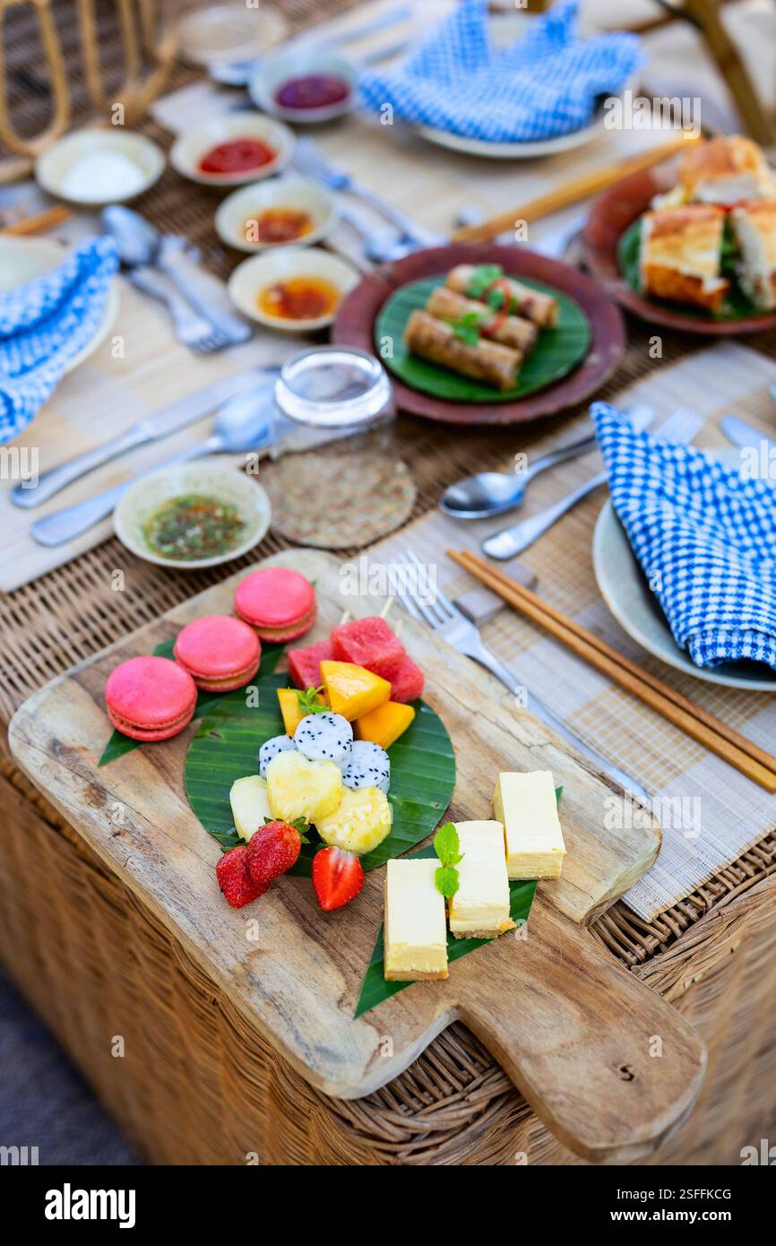 Un cadre de pique-nique magnifiquement aménagé avec une variété de desserts, y compris des macarons, des fruits tropicaux et des gâteaux, servis sur un plateau en bois Banque D'Images