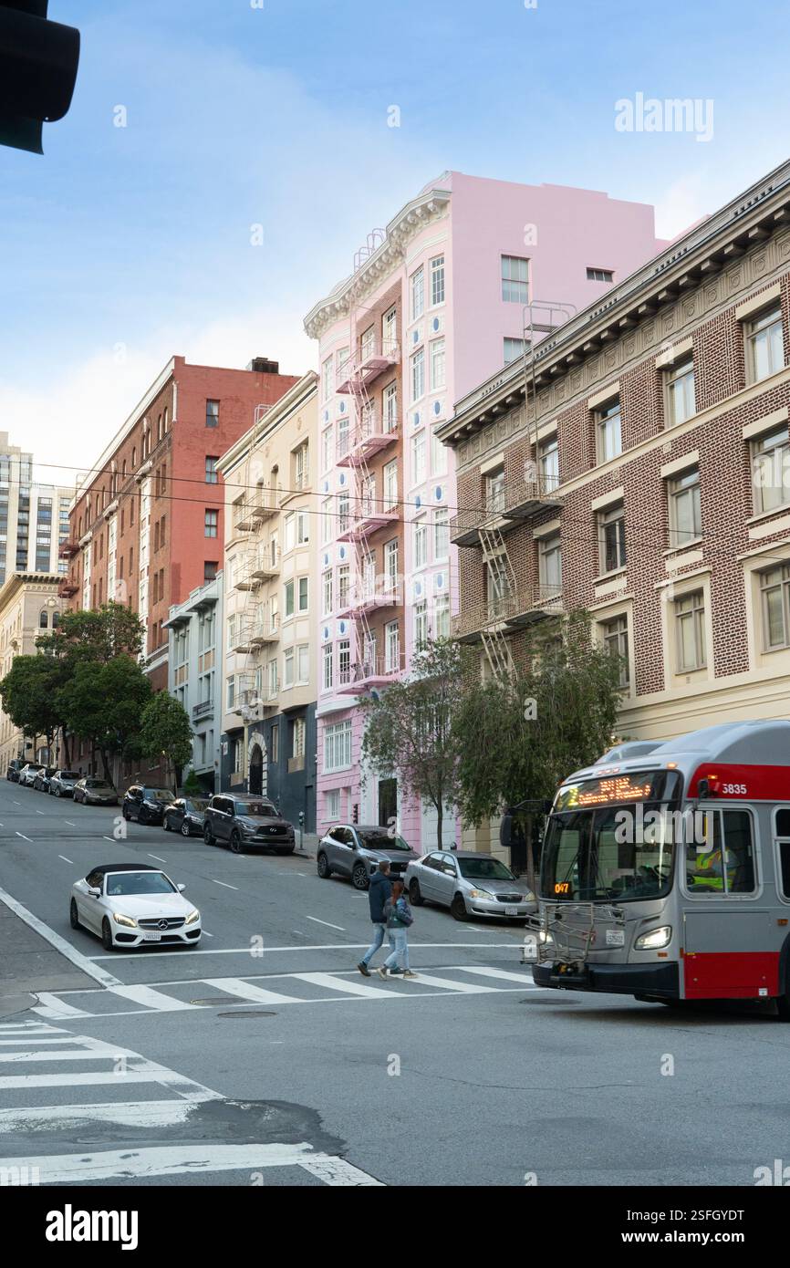 Les piétons traversent une rue de San Francisco, près des bâtiments emblématiques aux couleurs pastel sur une colline escarpée. Un bus muni passe. Banque D'Images