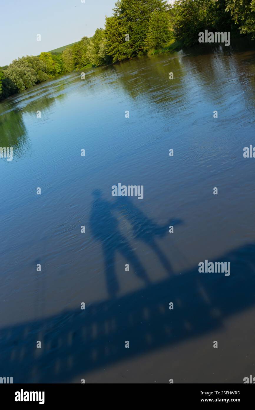 Deux silhouettes se tiennent près d'une rivière paisible, leurs ombres dansant à la surface de l'eau sous la lumière du soleil. Une végétation luxuriante entoure la scène, créa Banque D'Images
