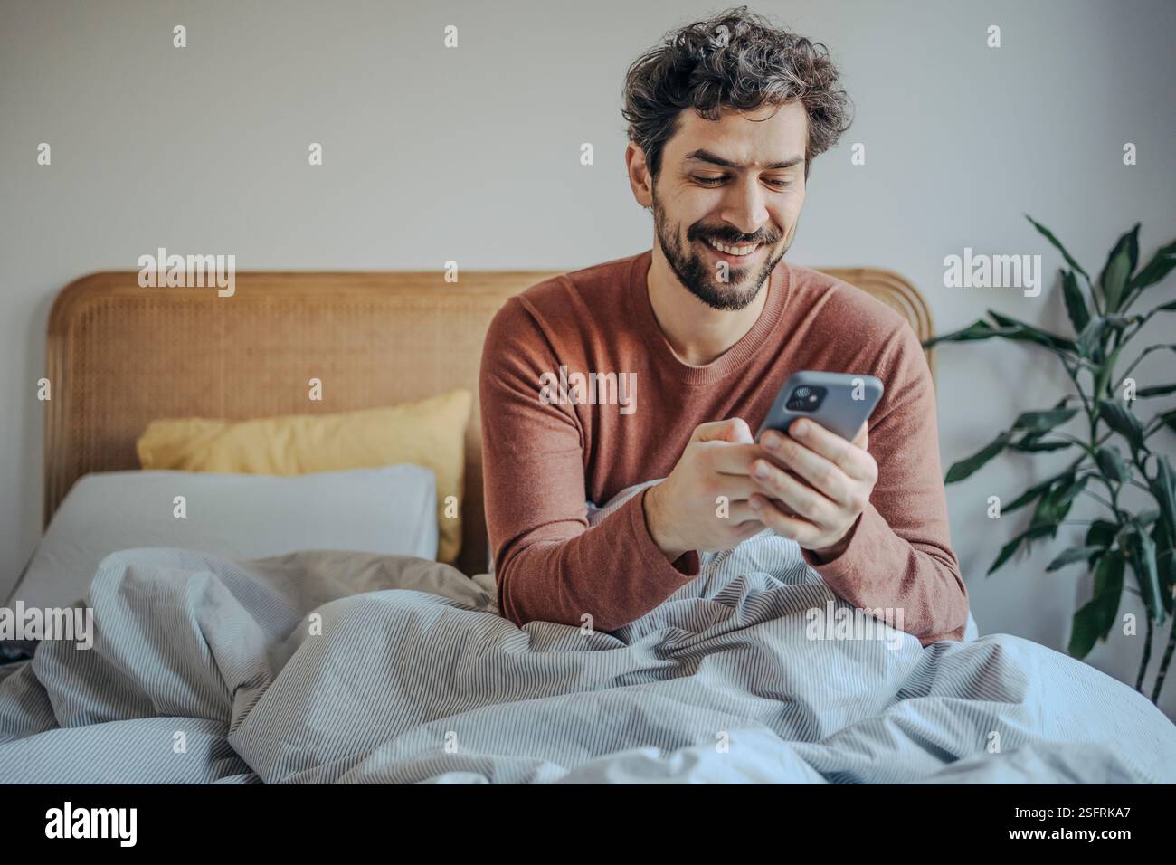 Routine matinale détendue – jeune homme barbu du moyen-Orient beau dans le lit vérifiant le téléphone. Banque D'Images