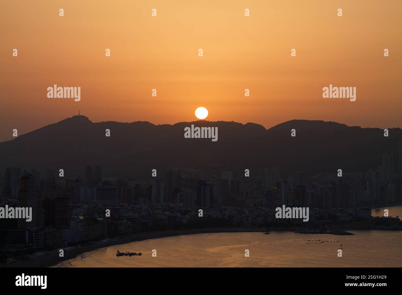Un coucher de soleil serein sur une ville côtière, avec le coucher de soleil derrière les montagnes. La ligne d'horizon comprend de hauts bâtiments et une plage calme, reflétant la chaleur d'oran Banque D'Images