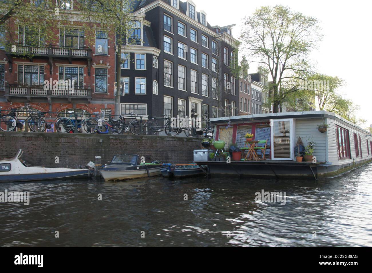 Une péniche en bois avec des fenêtres flotte sur un canal calme d'Amsterdam. Un bâtiment historique en brique avec de nombreuses fenêtres se dresse derrière la péniche, mettant en valeur th Banque D'Images