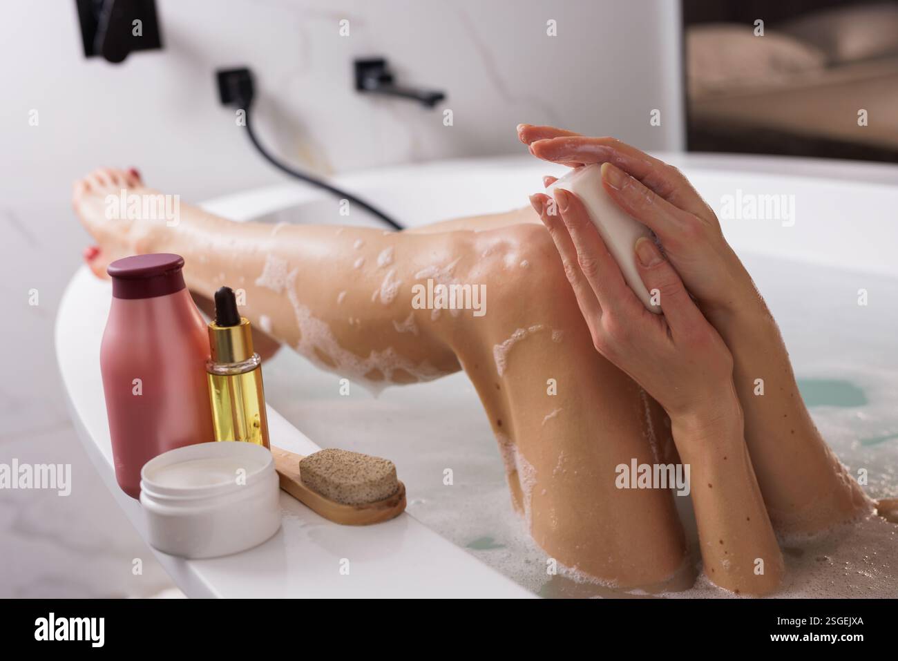 Temps de bain relaxant : femme profitant d'un rituel de soins personnels dans une baignoire, savon moussant sur ses mains. Les essentiels du spa comme une lotion pour le corps, de l'huile et une pierre ponce Banque D'Images