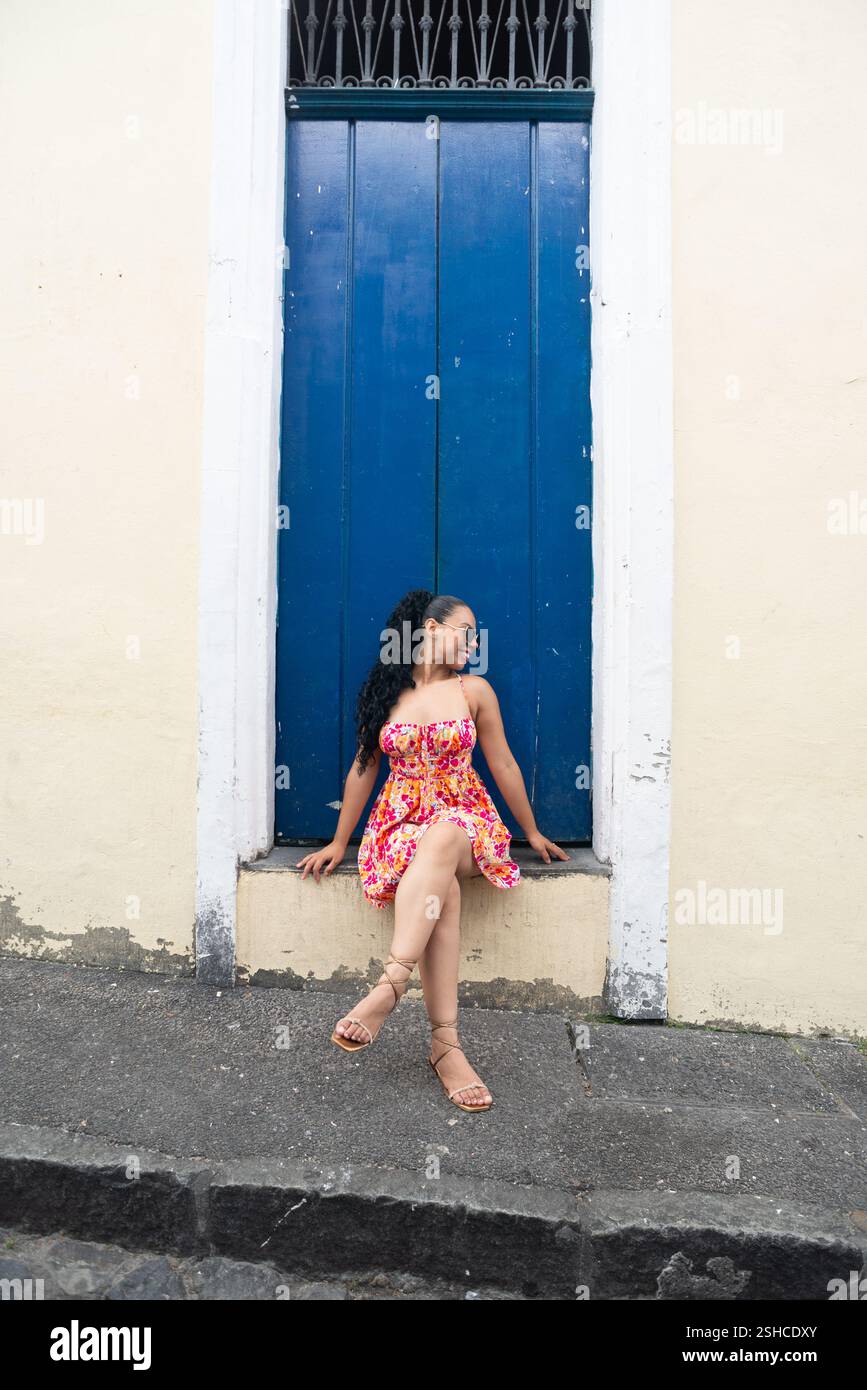 Belle jeune femme caucasienne avec des tresses et une robe colorée assise devant une porte bleue d'une vieille maison posant pour une photo. Personne confiante. B Banque D'Images