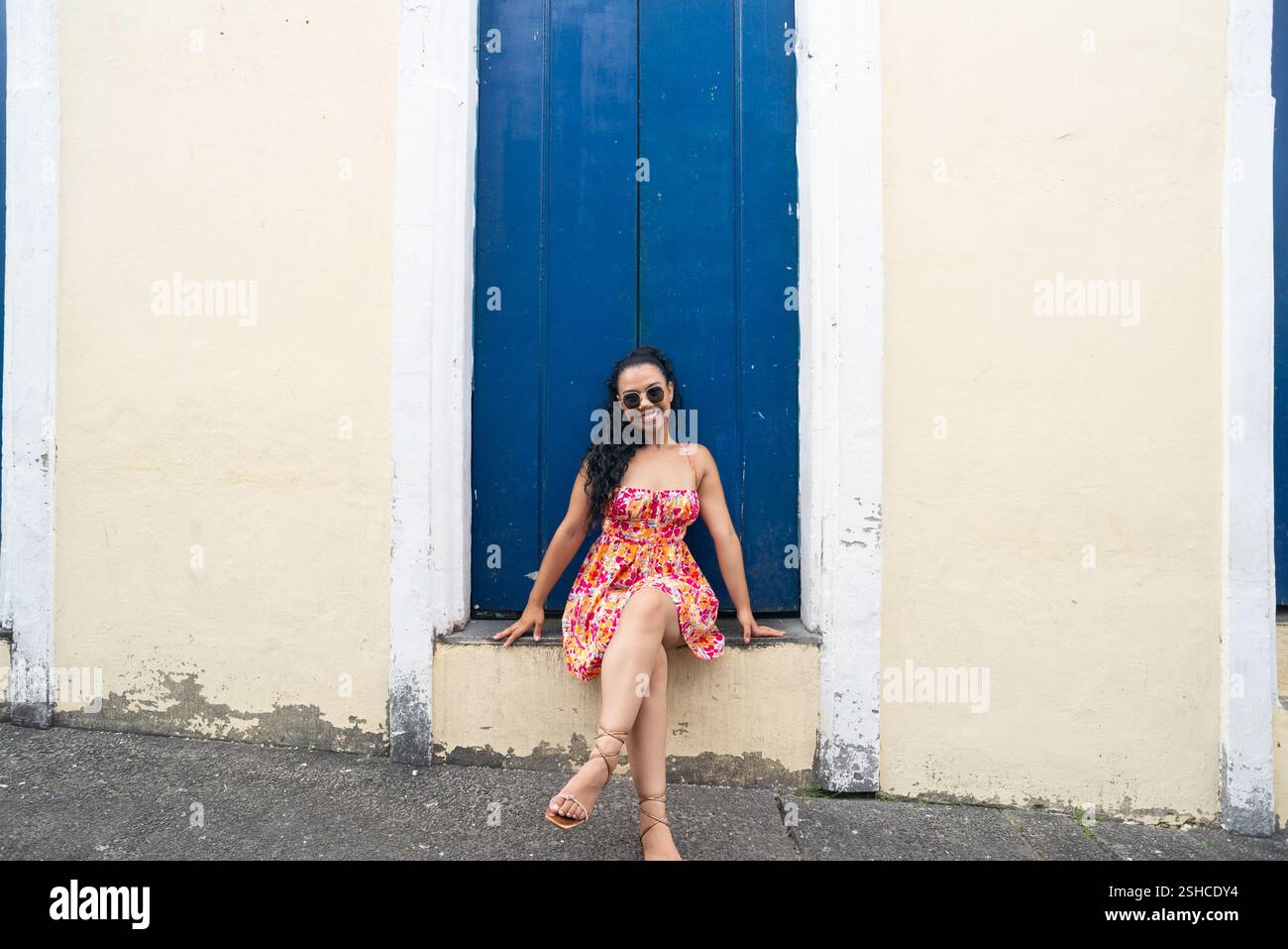 Belle jeune femme caucasienne avec des tresses et une robe colorée assise devant une porte bleue d'une vieille maison posant pour une photo. Personne confiante. B Banque D'Images