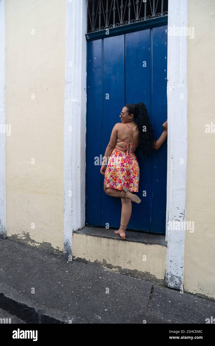 Belle jeune femme caucasienne avec des tresses, des lunettes de soleil et une robe colorée debout par une porte bleue d'une vieille maison. Personne confiante. Brésil Banque D'Images