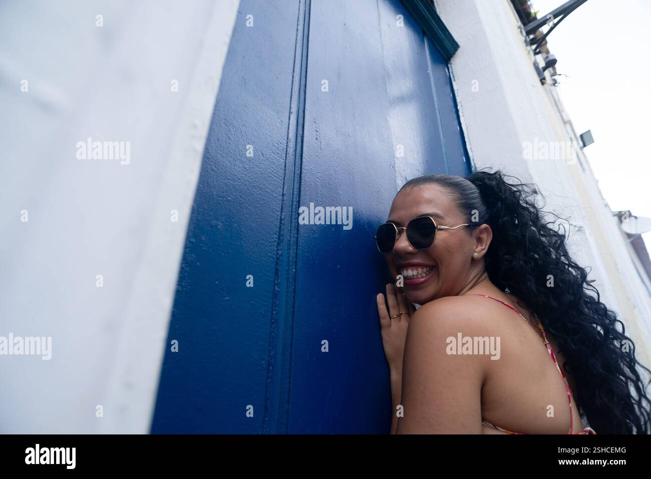 Belle jeune femme caucasienne avec des tresses, des lunettes de soleil et une robe colorée debout par une porte bleue d'une vieille maison. Personne confiante. Brésil Banque D'Images