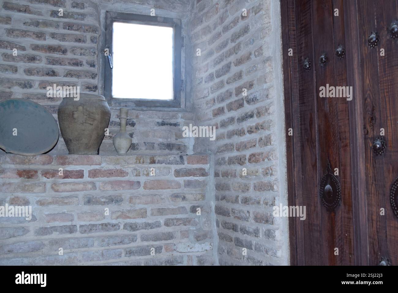 Intérieur rustique avec de la poterie ancienne sur une étagère en brique, avec une porte en bois avec des poteaux de fer traditionnels et une petite fenêtre, évoquant l'arc historique Banque D'Images