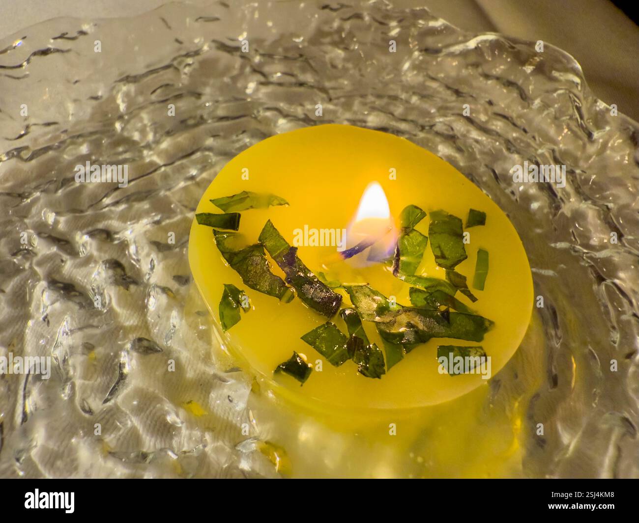 Bougie jaune ronde flottant dans l'eau dans un bol en verre texturé, brûlant avec des herbes sur le dessus Banque D'Images