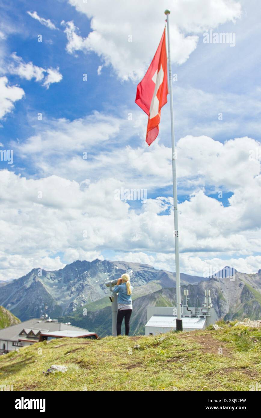 Une jeune fille utilisant des jumelles près d'un drapeau suisse, profitant d'une vue panoramique sur les Alpes en été, avec des pics montagneux spectaculaires et un ciel vibrant. Banque D'Images