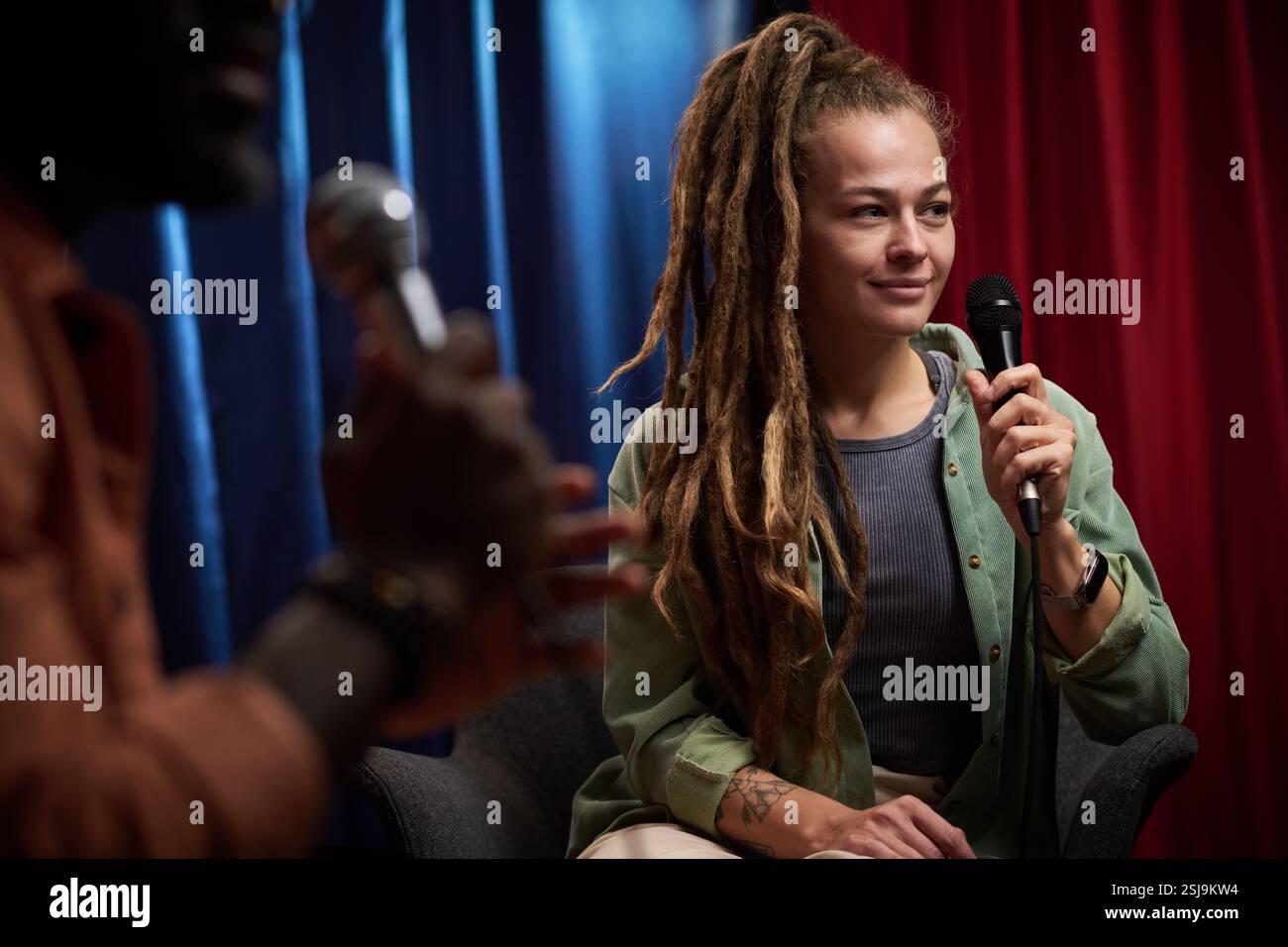 Femme avec de longs dreadlocks tenant le microphone tout en étant assis sur le panneau. S'engager dans une discussion réfléchie avec des rideaux colorés en arrière-plan Banque D'Images