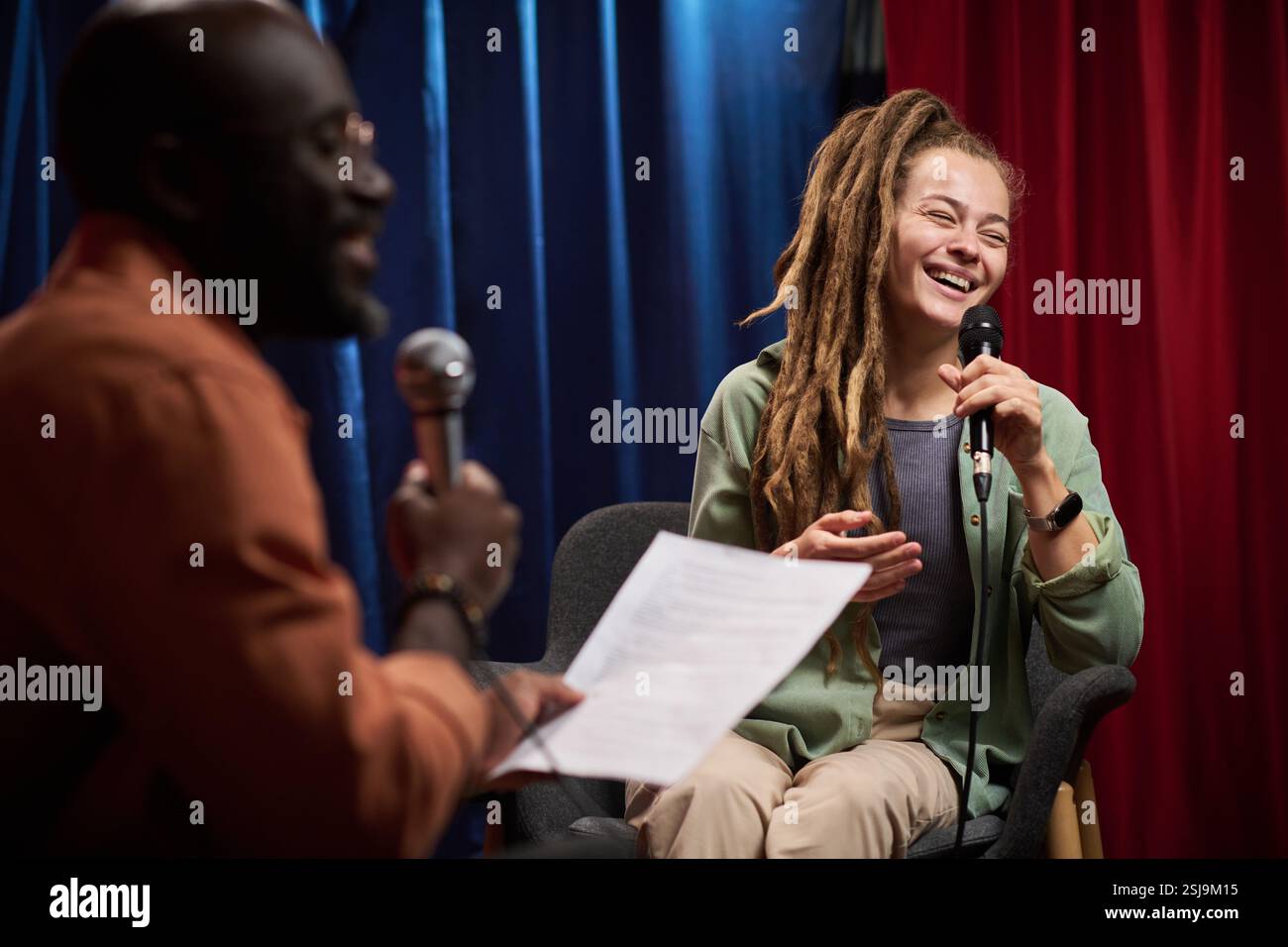 Jeune femme avec de longs dreadlocks riant tout en participant à un talk-show avec intervieweur dans une atmosphère détendue sur scène avec des rideaux bleus et rouges en arrière-plan Banque D'Images
