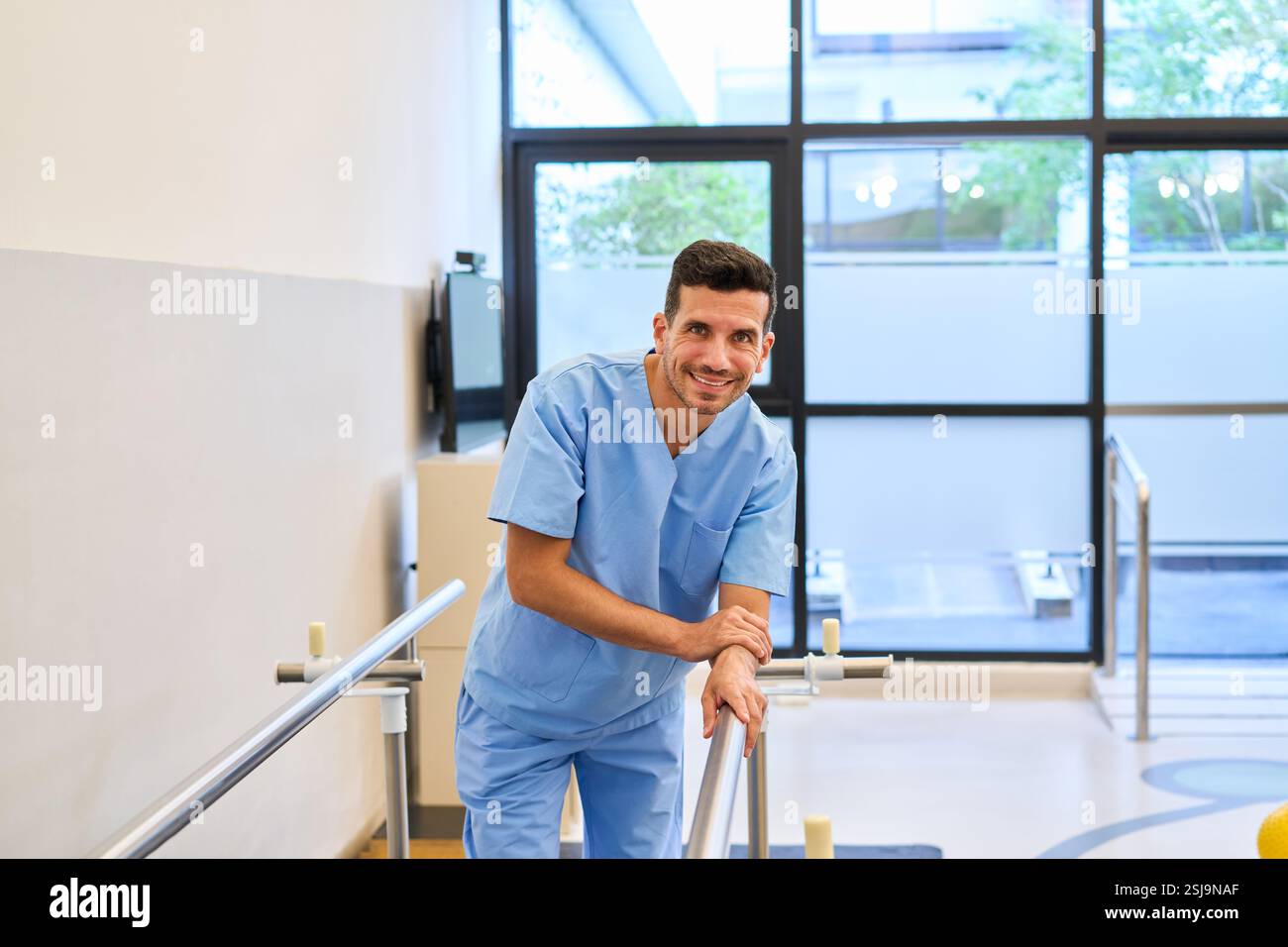 Travailleur de la santé masculin souriant tout en se tenant debout près de l'équipement de physiothérapie dans un centre de réadaptation. L'environnement met l'accent sur la santé, la réhabilitation Banque D'Images