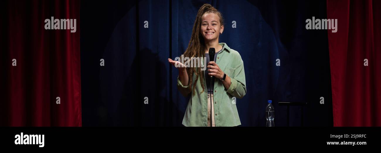 Femme debout sur scène souriante tout en tenant le microphone à la main partageant un moment joyeux pendant la performance sur un fond sombre Banque D'Images
