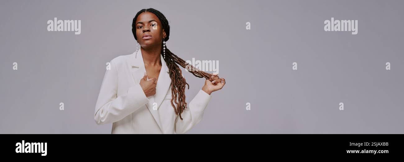 Portrait de femme élégante avec de longues tresses et veste blanche décontractée tenant ses cheveux. L'arrière-plan est minimal et gris, attirant l'attention sur sa position serrée et la texture de ses tresses Banque D'Images