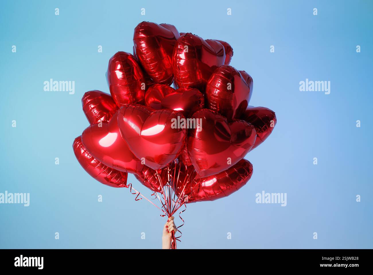 Main tenant un tas de ballons rouges en forme de cœur contre un ciel bleu vibrant, symbolisant l'amour, la célébration et la joie. Banque D'Images