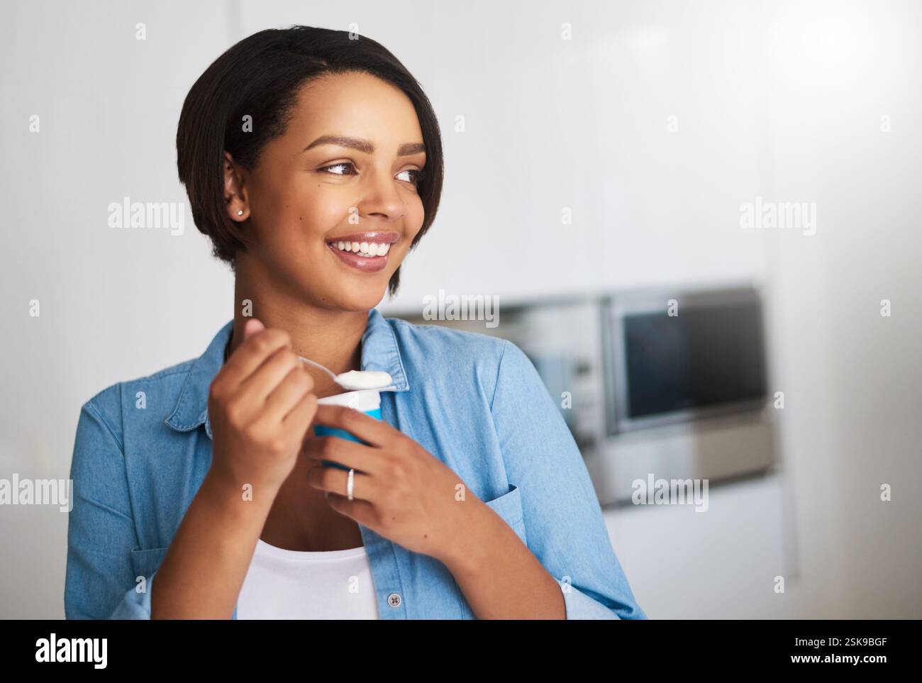 Femme, heureuse et pensant avec du yaourt dans la cuisine, le petit déjeuner et la réflexion avec collation laitière. Personne, fille et affamée avec le sourire, la nutrition et l'équilibre Banque D'Images