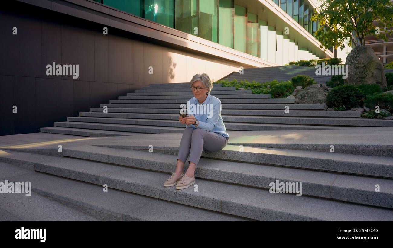 Réfléchie femme d'affaires mature aux cheveux gris des années 50 femme caucasienne senior femme tenir le téléphone pensant taper le message mobile chat messagerie entreprise mondiale Banque D'Images