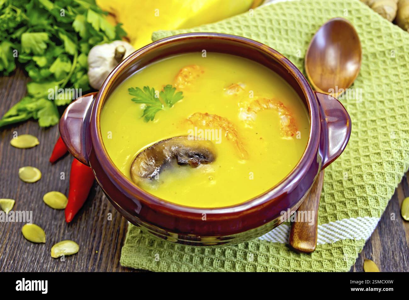 Soupe-purée citrouille avec crevettes et champignons dans un bol en argile avec une cuillère sur la serviette, citrouille, persil, gingembre, gousses de piment, graines de citrouille sur t Banque D'Images