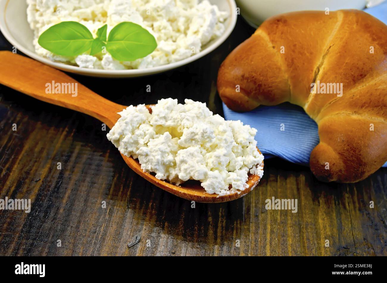 Le fromage cottage dans une soucoupe avec du basilic sur une cuillère en bois, un bagel, une cruche de lait, nappes sur planche de bois Banque D'Images