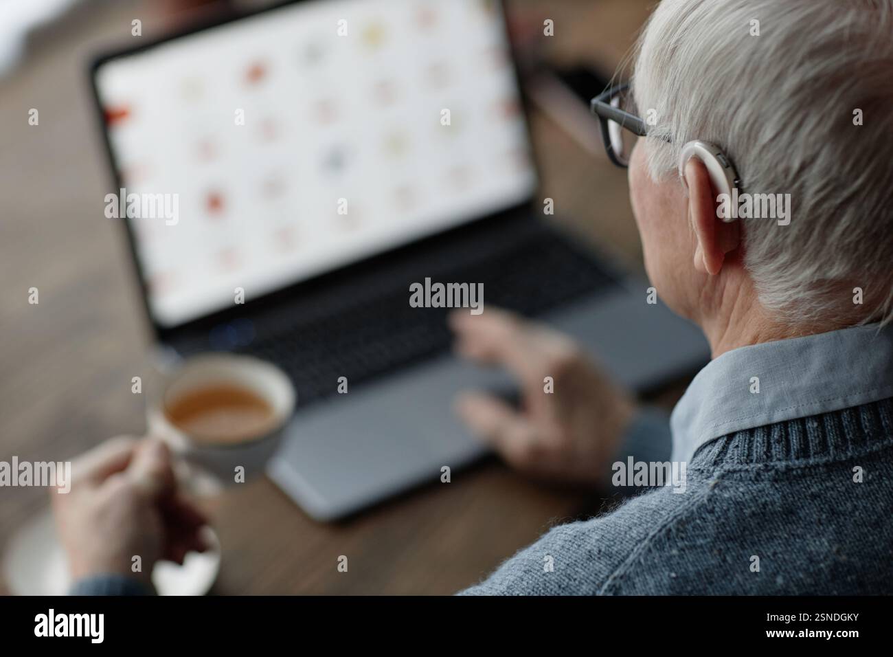 Photo au-dessus de l'épaule d'un homme âgé portant une prothèse auditive derrière l'oreille travaillant sur un ordinateur portable à la maison tout en utilisant une technologie d'assistance, espace de copie Banque D'Images
