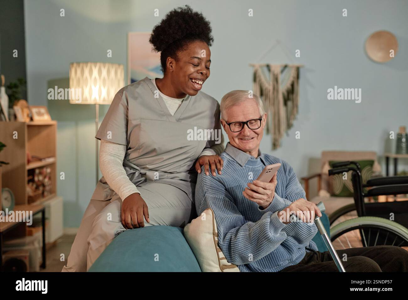 Cliché franc d'une travailleuse sociale noire souriante aidant un homme âgé à utiliser la technologie tout en regardant une vidéo au téléphone ensemble dans le salon Banque D'Images