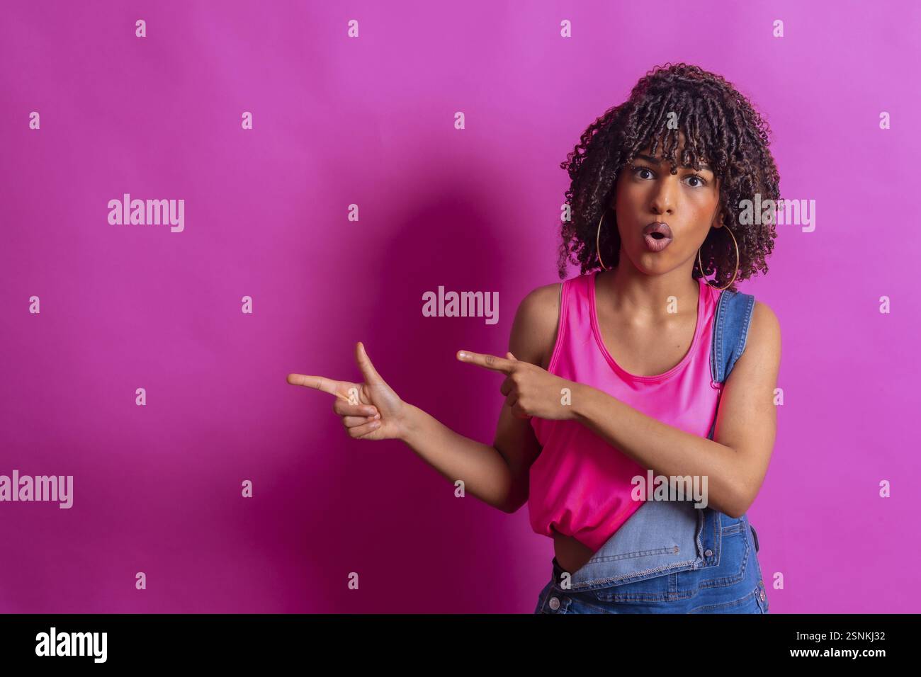 Jeune femme surprise avec des pointes de cheveux bouclés sur le côté avec une expression choquée, suggérant une opportunité promotionnelle contre un backdr rose vibrant Banque D'Images