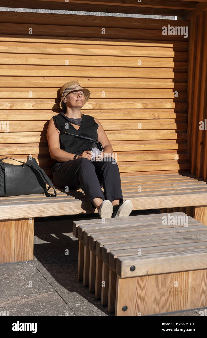 Femme élégante avec chapeau de paille assis sur un banc en bois et se détendre dans une journée d'été ensoleillée à Andermatt, Uri, Suisse, Europe Banque D'Images