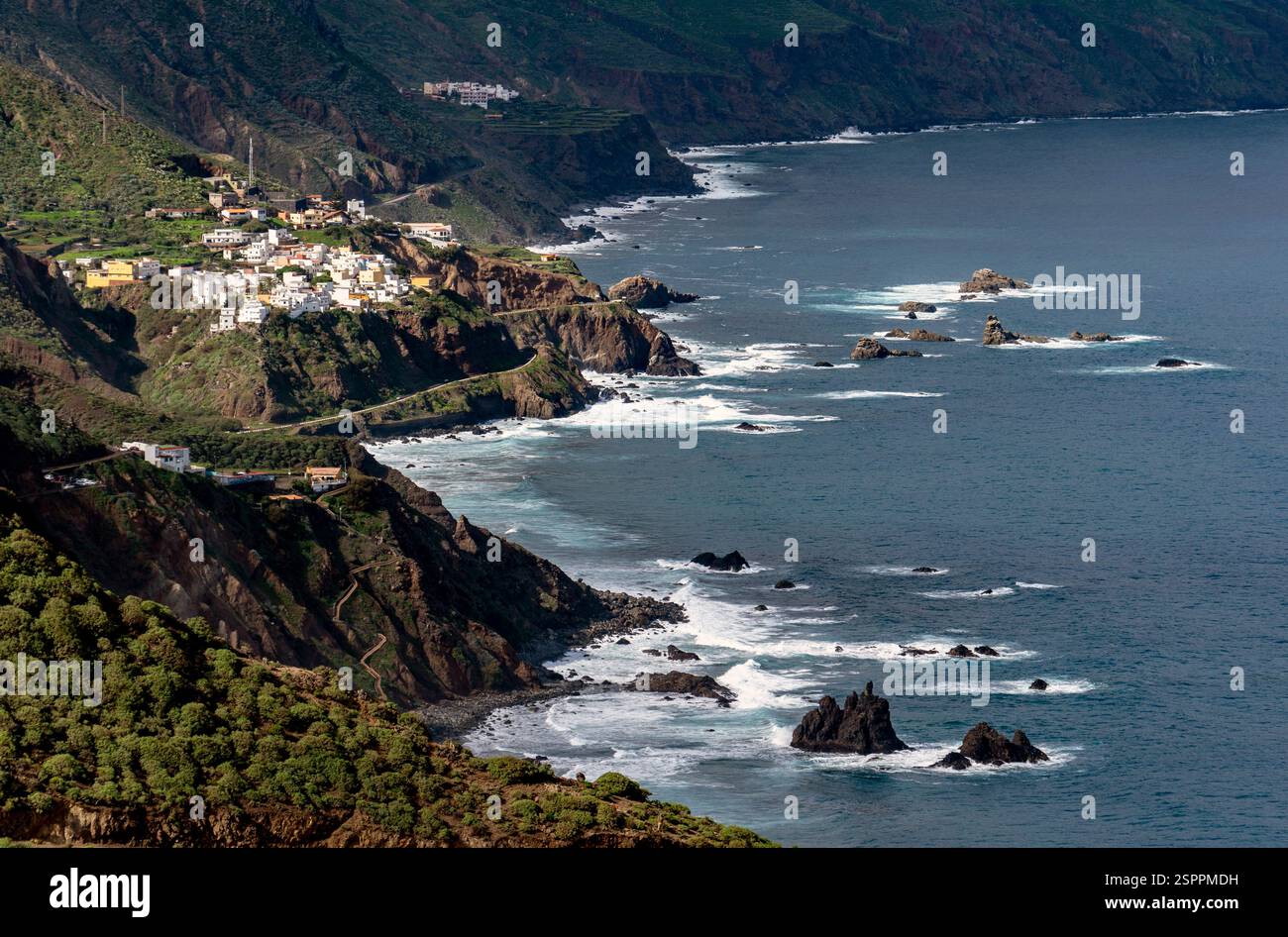 Une vue panoramique sur la côte avec des falaises accidentées, des vagues qui s'écrasent contre des rochers et un petit village perché sur la colline. Le paysage est luxuriant Banque D'Images