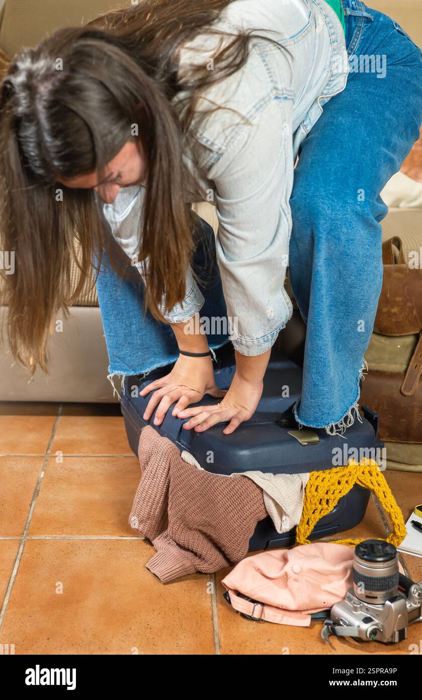 Une jeune femme se tient debout sur sa valise rembourrée, dans une pose amusée et avec une expression d'effort, pressant ses mains ensemble dans une tentative de clo Banque D'Images